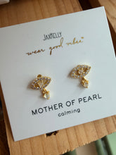 Load image into Gallery viewer, A pair of gold-toned eye-shaped stud earrings with clear rhinestones and a small clear pearl drop, displayed on a white Jaxxelly brand card labeled 'MOTHER OF PEARL calming' on a wooden surface.